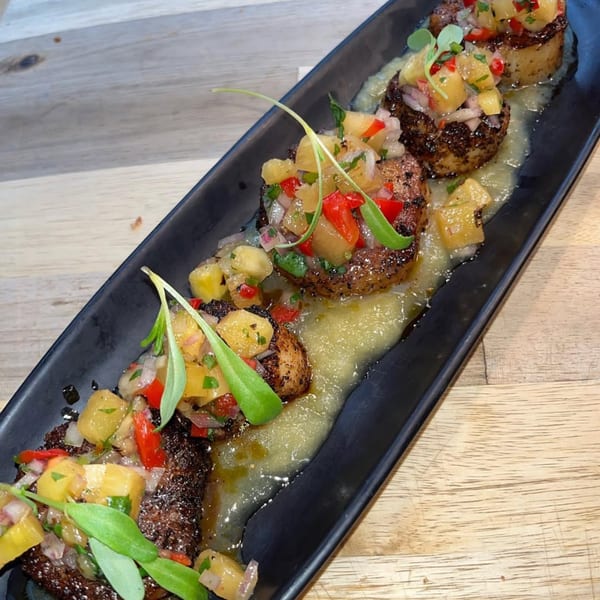 Grilled steak medallions topped with pineapple salsa and microgreens, served on a black rectangular plate.