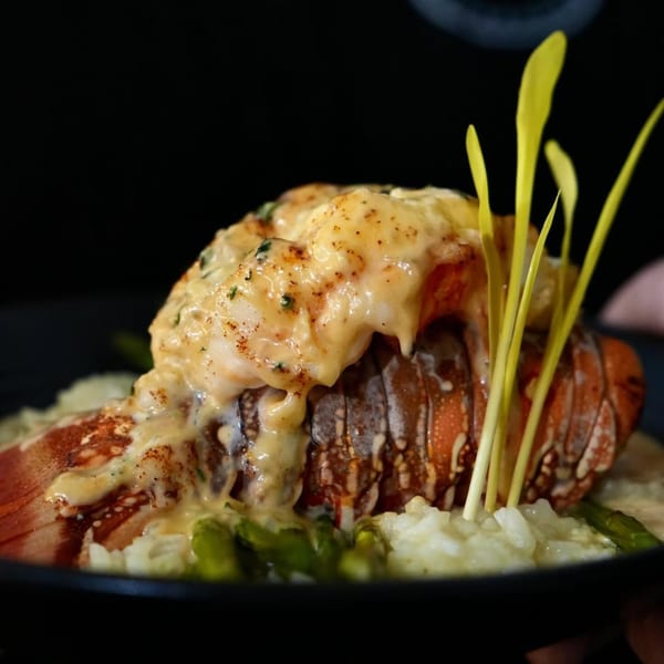 Lobster tail with creamy sauce, risotto, asparagus, and green garnish on a dark plate.