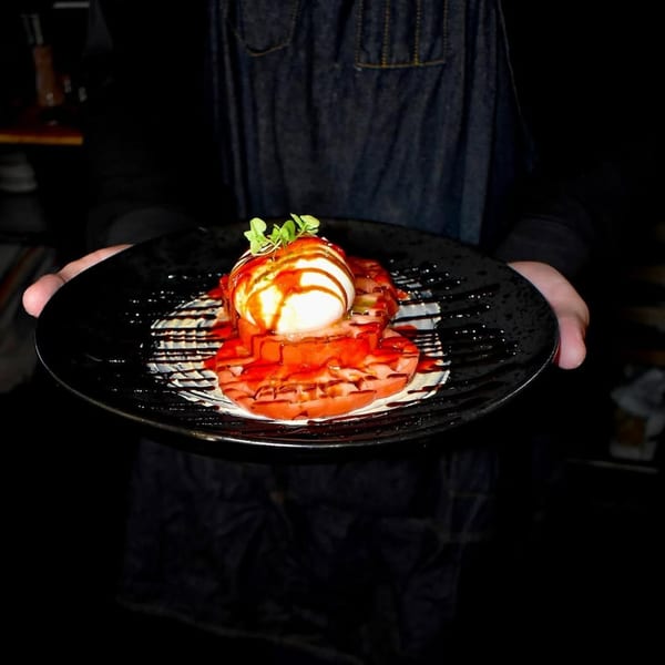 Hands holding a black plate with sliced tomatoes, burrata, balsamic glaze, and microgreens.