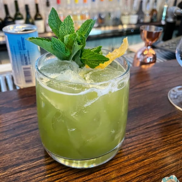 A vibrant green cocktail with mint and a lemon peel garnish sits on a wooden bar.