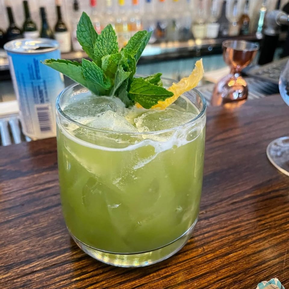 A vibrant green cocktail with mint and a lemon peel garnish sits on a wooden bar.