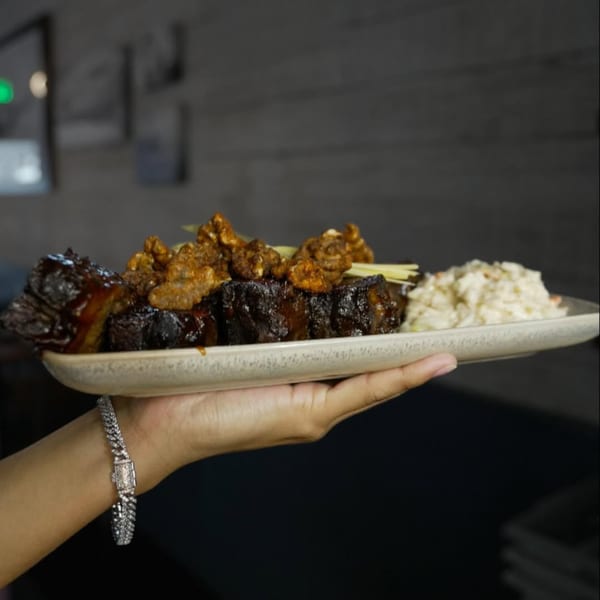 A hand holding a plate of Berkeley Cut Steakhouse's glazed ribs, crispy pork belly, and coleslaw.