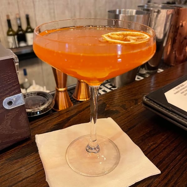 A vibrant orange cocktail with a dried orange slice, on a wooden bar at Berkeley Cut Steakhouse.