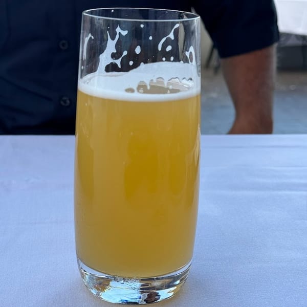 A tall glass of golden beer with a foamy head sits on a white tablecloth.