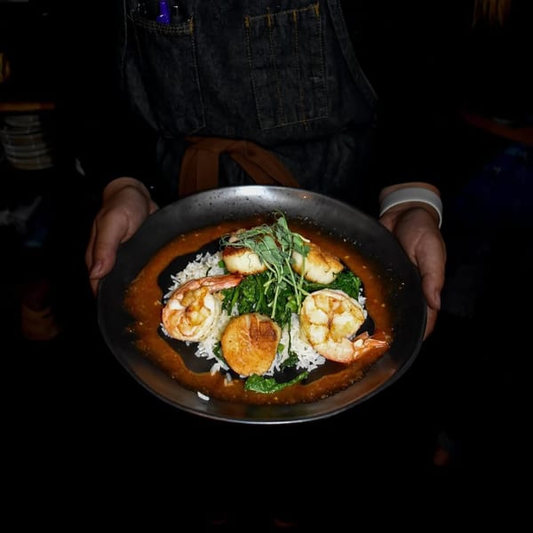 A server presents a dish with shrimp, scallops, rice, and greens on a dark plate.